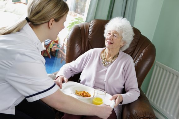 Une aide-soignante sert un repas à une personne âgée assise dans un fauteuil.